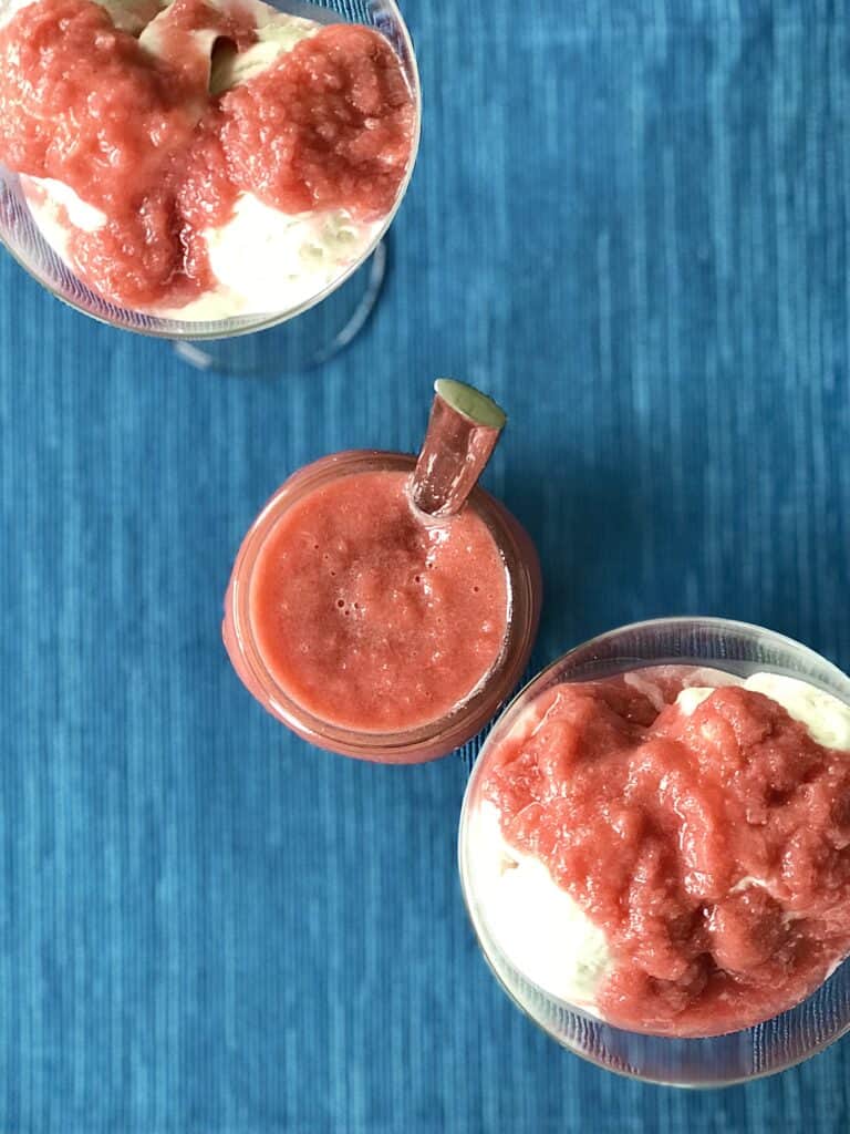 healthy rhubarb sauce in a mason jar with a spoon and in two coupe glasses with vanilla ice cream, all on top of a blue table runner