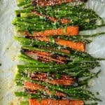 carrots and asparagus roasted in the oven, coated in sesame seeds, sprinkled with chopped parsley, on a foil-lined baking sheet on a wooden table