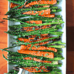 roasted asparagus and carrots sprinkled with sesame seeds and chopped parsley on a white platter with silver tongs