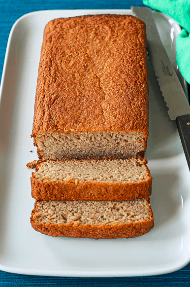 gluten-free banana bread sliced on a white platter with a serrated knife and green towel, sitting on a blue table runner