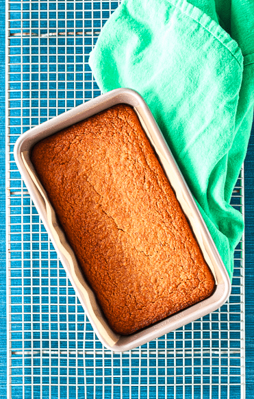 healthy banana bread in a metal loaf pan on a cooling rack next to a green towel, on a blue table runner
