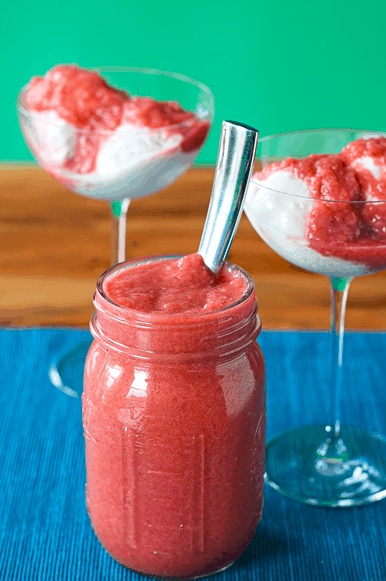 a sauce made of rhubarb, honey and vanilla fills a mason jar with a spoon and is poured over vanilla ice cream in two coupe glasses, all on a blue table runner on a wooden table