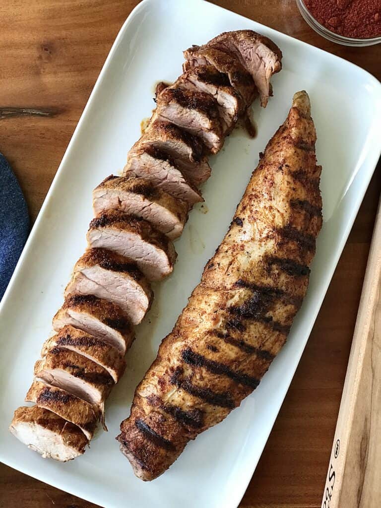 2 pork tenderloins with char marks on a white platter - one whole, one sliced