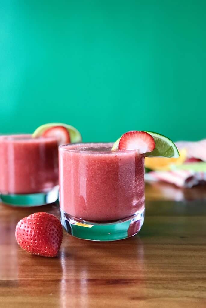 a healthy frozen mocktail made with strawberries in short glasses on a wooden table strewn with strawberries, orange slices and lime slices