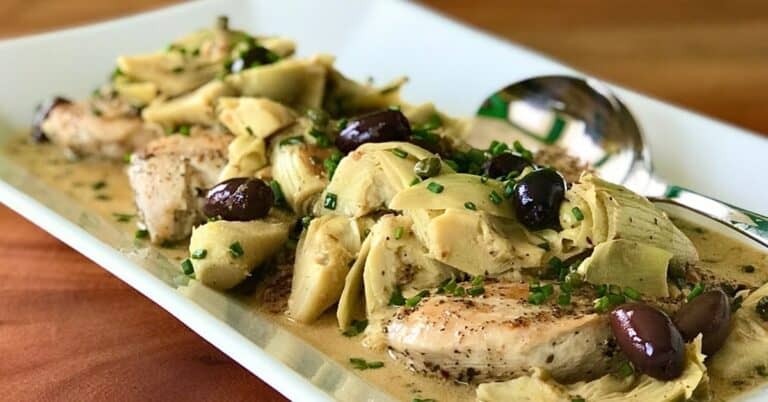 Paleo lemon chicken with olives, artichokes, capers and chives on a white platter with a large silver serving spoon