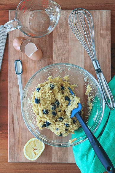 the raw batter for a gluten-free blueberry cake in a glass bowl on a wooden cutting board with egg shells, a measuring spoon, a measuring glass and half a squeezed lemon