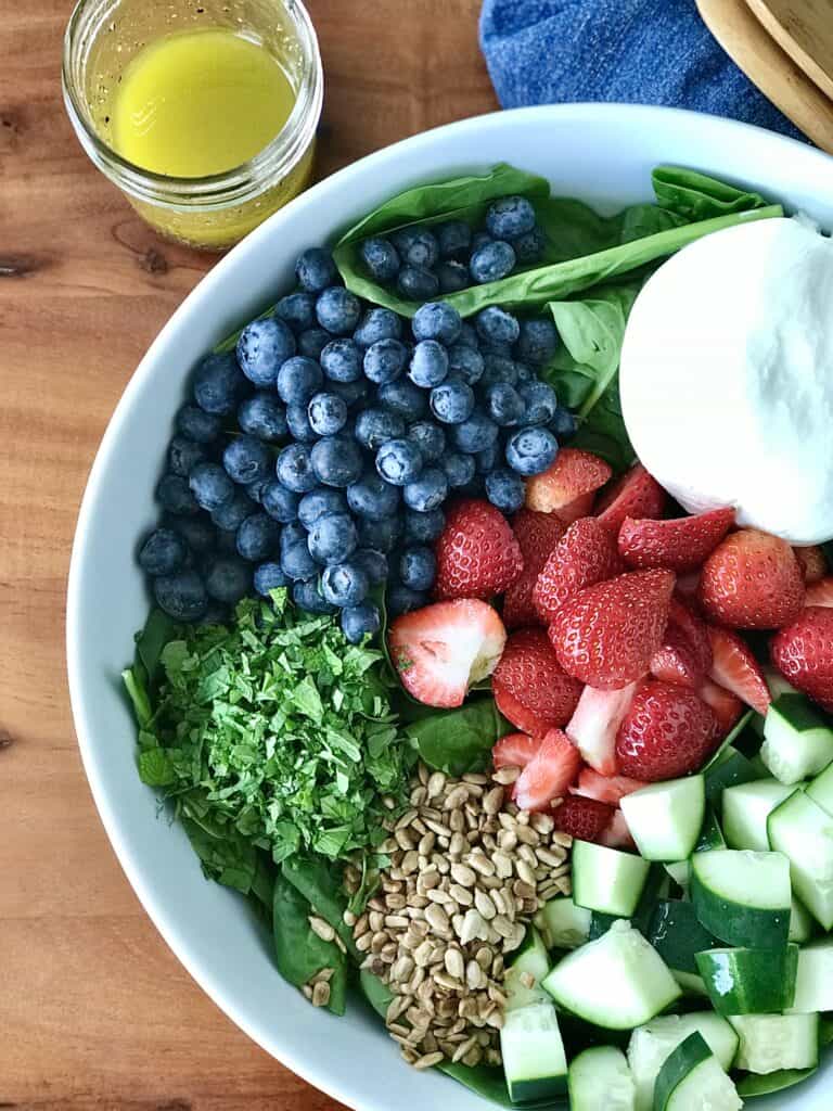 a white bowl filled with spinach, berries, burrata, cucumber, sunflower seeds and herbs