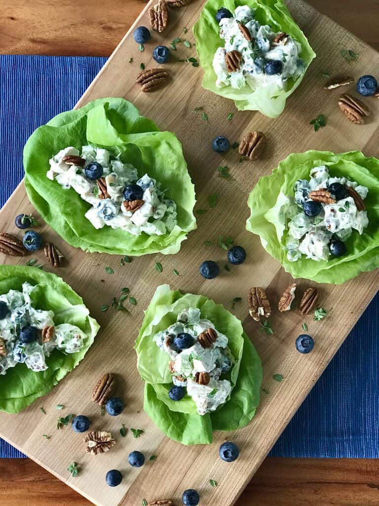 Paleo chicken salad piled into lettuce cups on a wooden cutting board sprinkled with blueberries, pecans and fresh thyme leaves