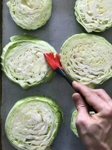 cabbage slices on a baking sheet getting brushed with oil by a hand holding a black and white silicone basting brush
