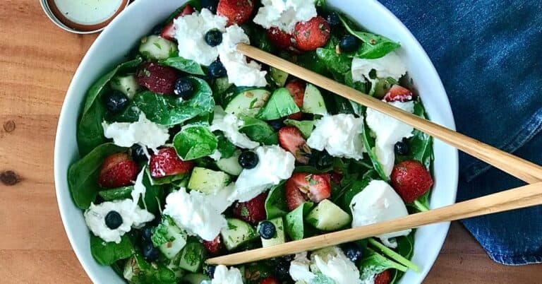 a berry spinach salad with burrata and cucumber in a white bowl with wooden tongs, next to a blue towel on a wooden table