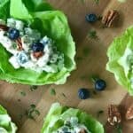 healthy chicken salad piled into lettuce cups on a wooden cutting board strewn with blueberries, pecans and fresh thyme leaves