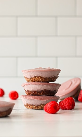 healthy raspberry cheesecake bites stacked on top of each other next to more cheesecake bites and fresh raspberries