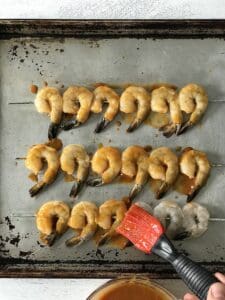 a silicone brush brushing BBQ sauce onto shrimp skewers resting on a baking sheet
