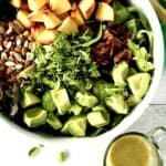 a big white bowl with a peach salad piled with leafy greens, almonds, avocado and basil with the dressing in a glass pitcher on the side