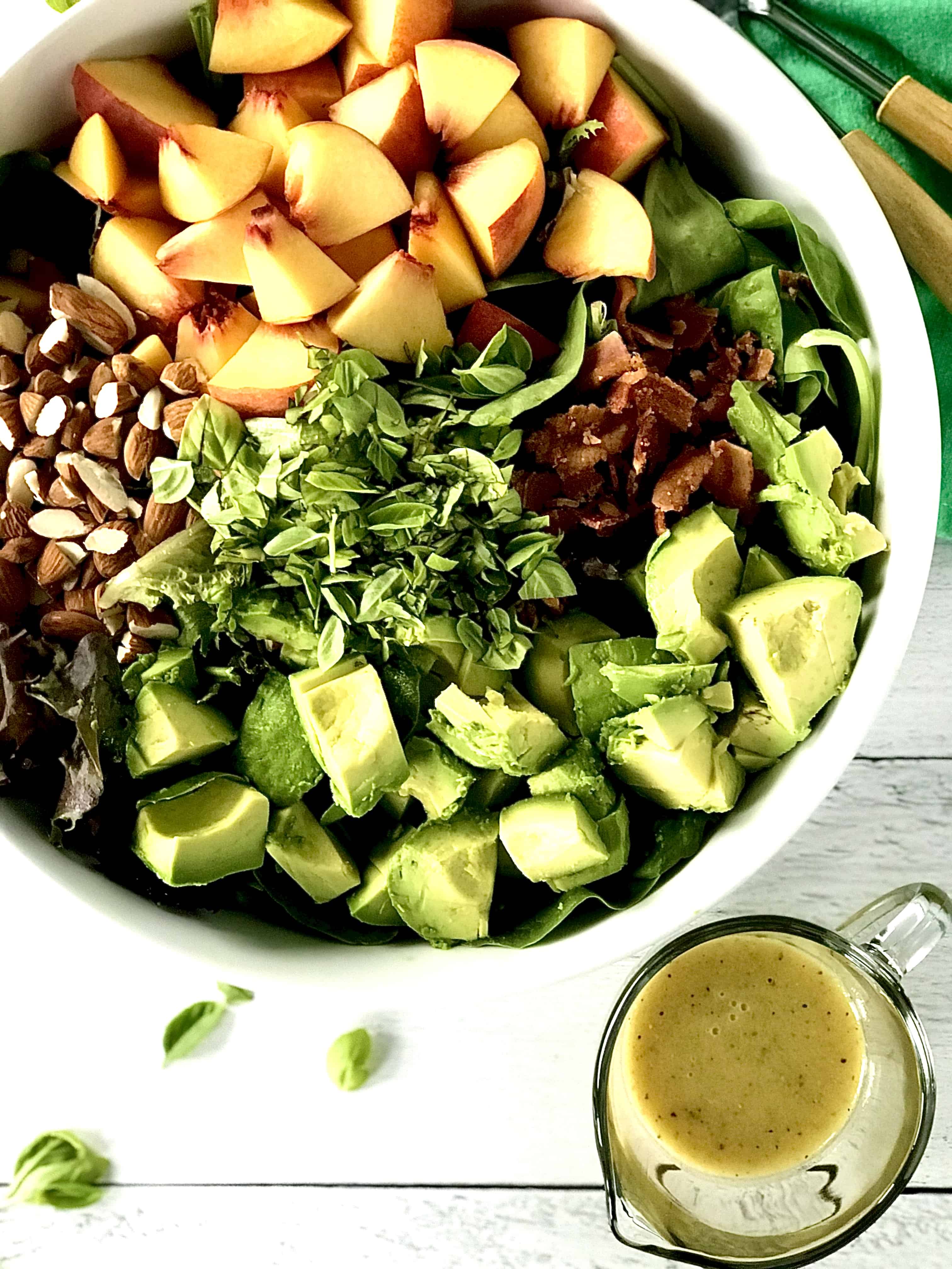 a big white bowl with a peach salad piled with leafy greens, almonds, avocado and basil with the dressing in a glass pitcher on the side