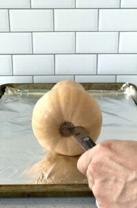 A hand sticking a paring knife into the bottom of a butternut squash on a baking sheet