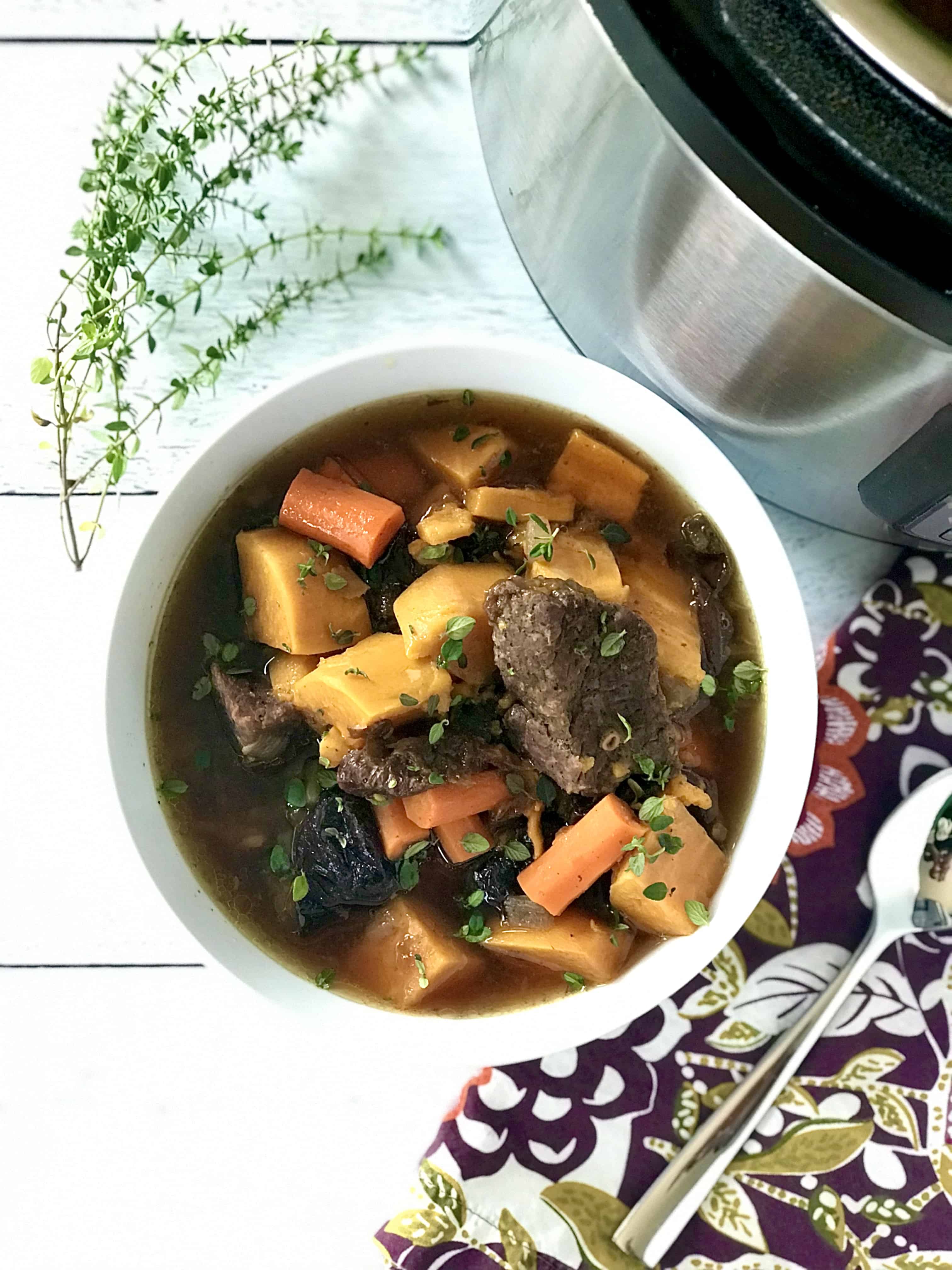 beef stew with root vegetables in a white bowl on a wooden table next to a floral napkin, an Instant Pot and a sprig of fresh thyme