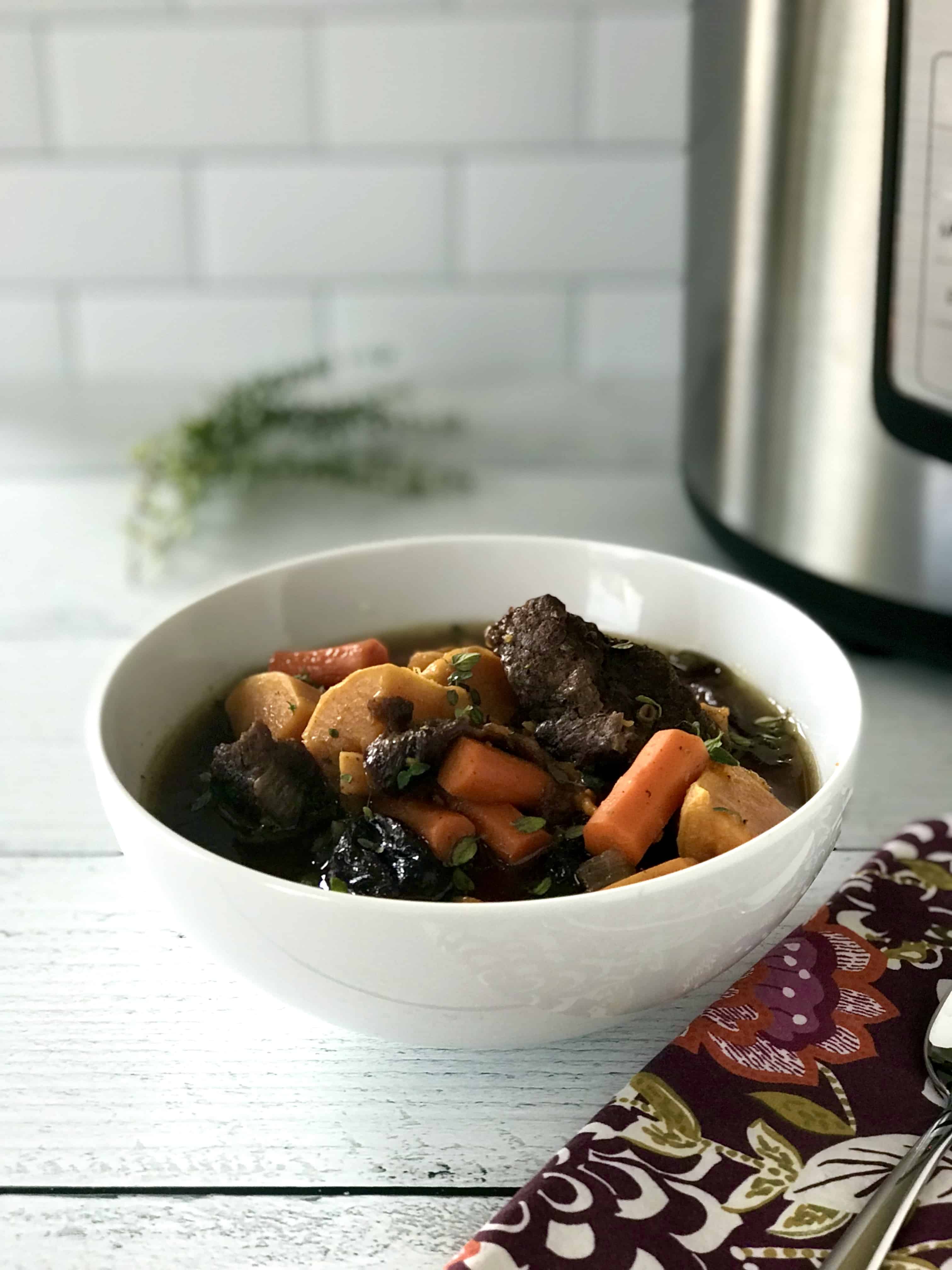 a root vegetable stew with chunks of beef in a white bowl on a white wooden table next to an Instant Pot and a sprig of fresh thyme