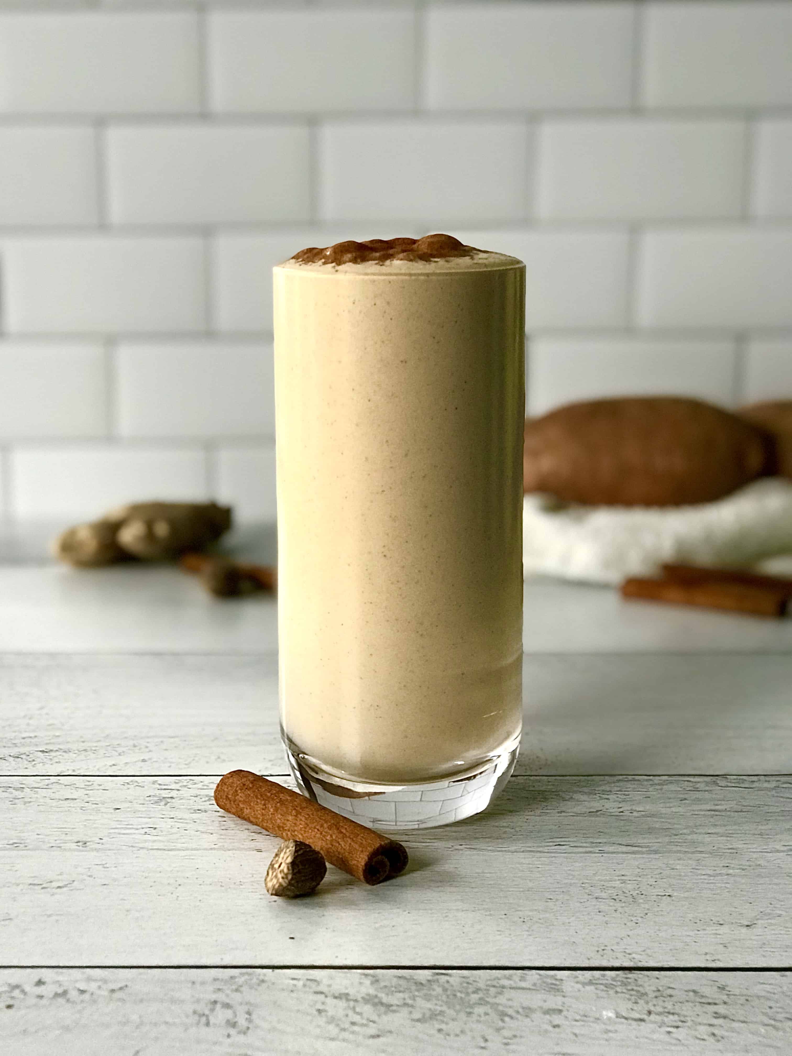 a protein smoothie made with sweet potato in a tall glass next to a cinnamon stick and half a nutmeg
