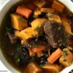 a stew with root vegetables and chunks of beef in a white bowl on a white wooden table