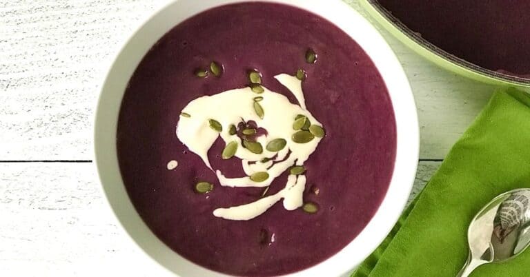 a blended purple sweet potato soup in a white bowl with a white cream drizzle and pumpkin seeds on top