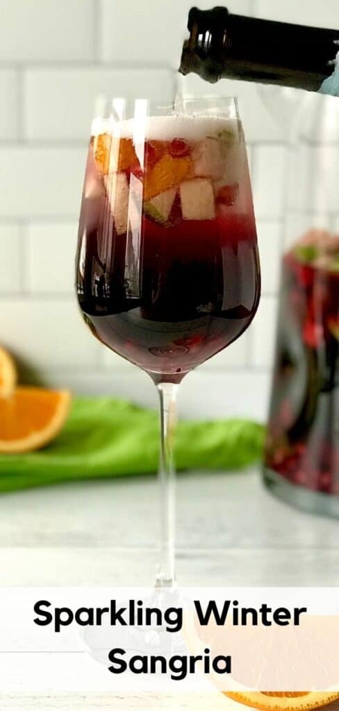 a bottle of Prosecco pouring Prosecco into a wine glass full of Sparkling Winter Sangria with apples, oranges and pomegranate seeds