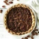 a dairy-free pecan pie in a pie dish on a white wooden table surrounded by pecans