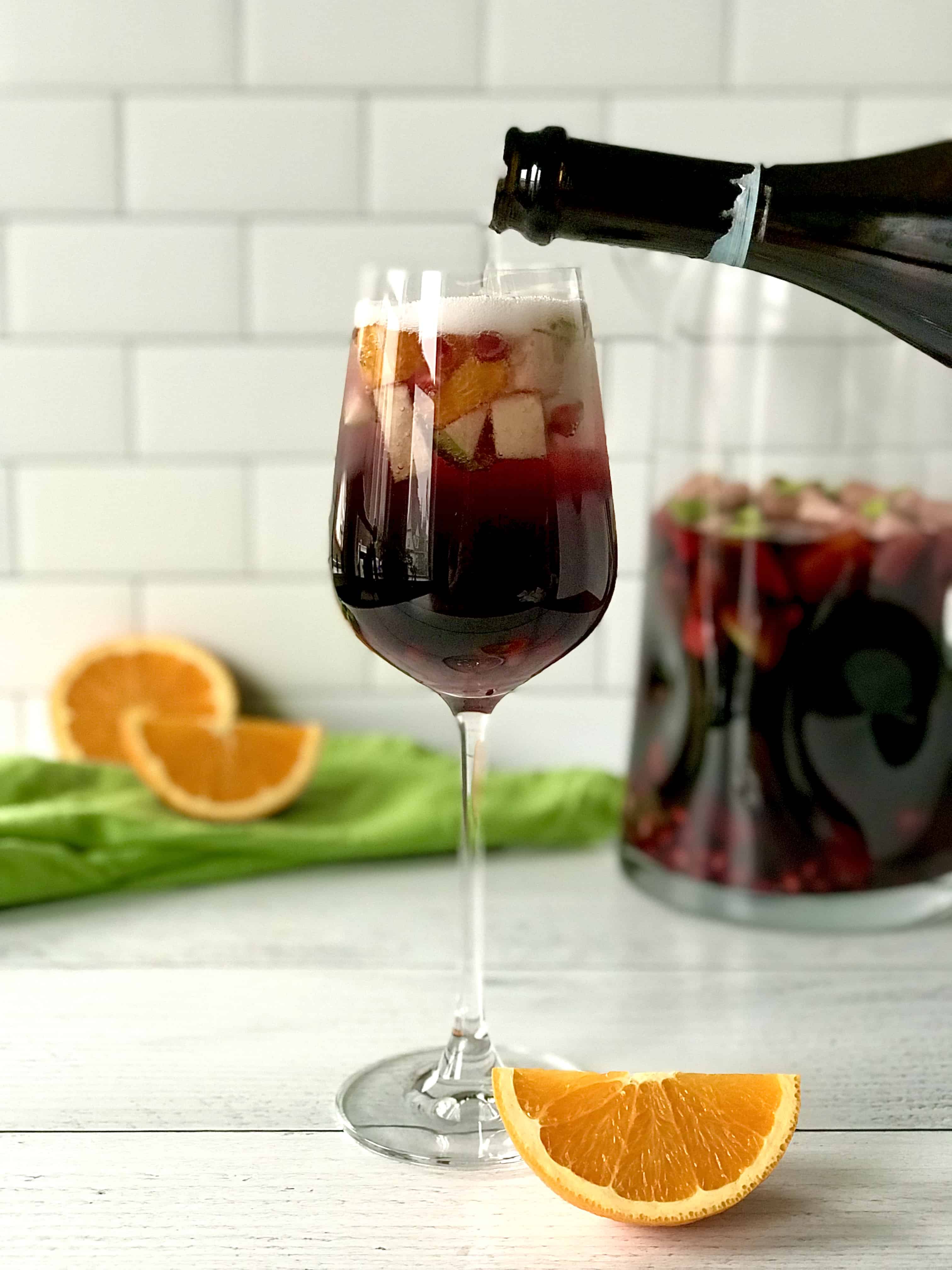 a wine glass full of red wine sangria with apples, oranges and pomegranate seeds floating in it, with a bottle of Prosecco pouring bubbly wine on top