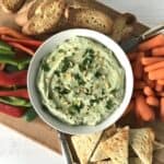 a ricotta dip blended with lemon, parsley and anchovies in a white bowl on a wooden cutting board with bell pepper strips, baby carrots and sliced toasted bread and baguettes