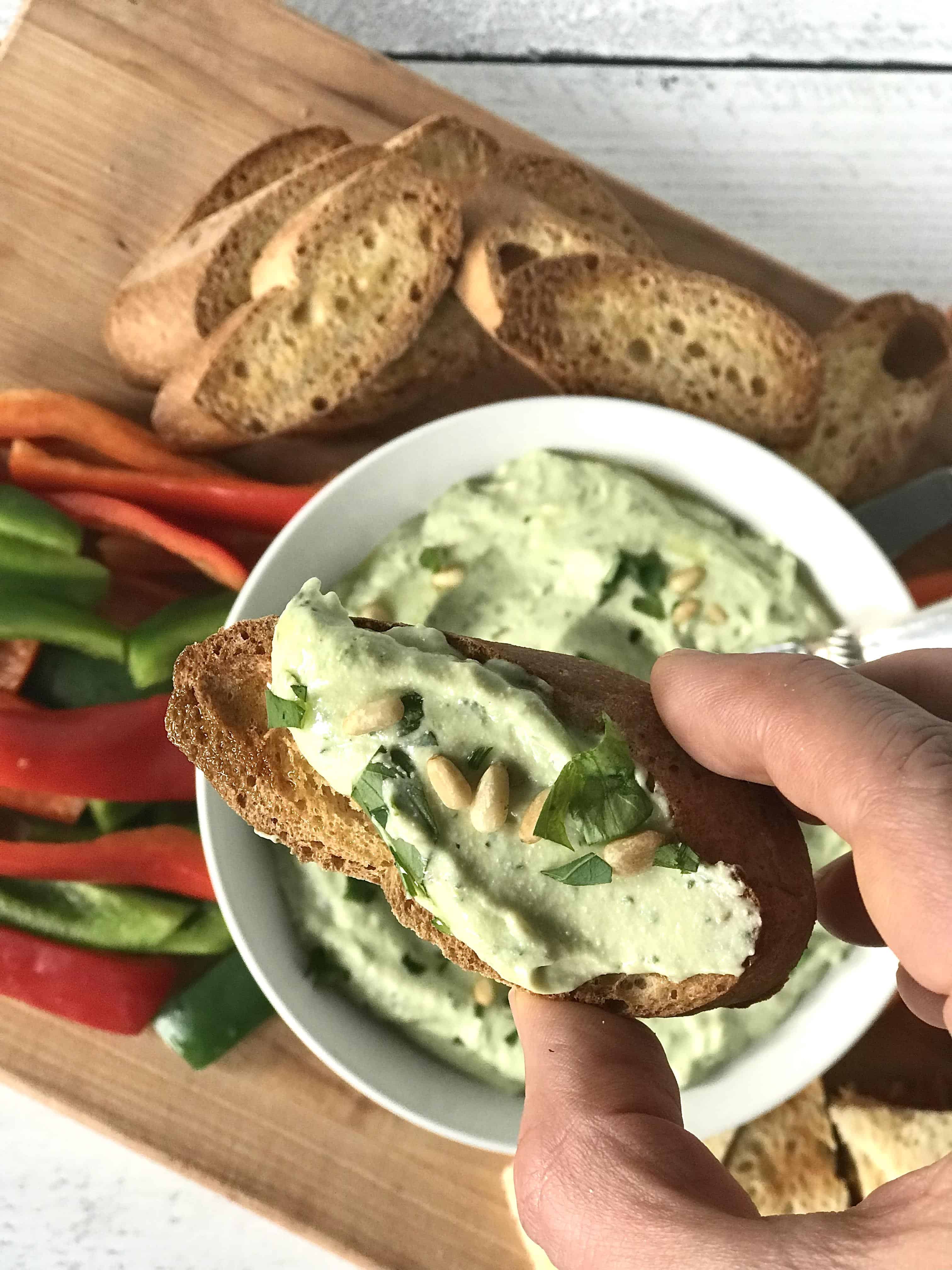 a creamy herb dip spread on a toasted baguette slice being held up by a hand