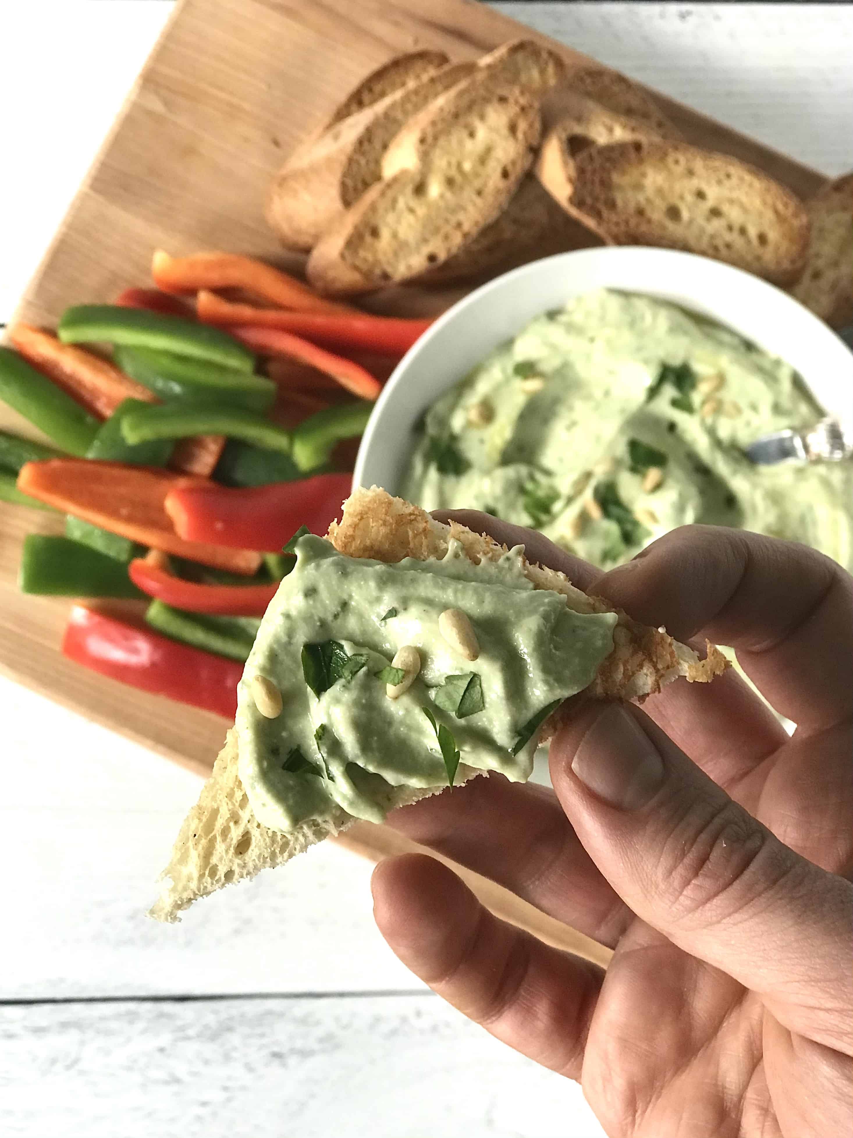 a hand holding up a toasted slice of bread cut into a triangle spread with a creamy pale green dip and topped with pine nuts and chopped parsley
