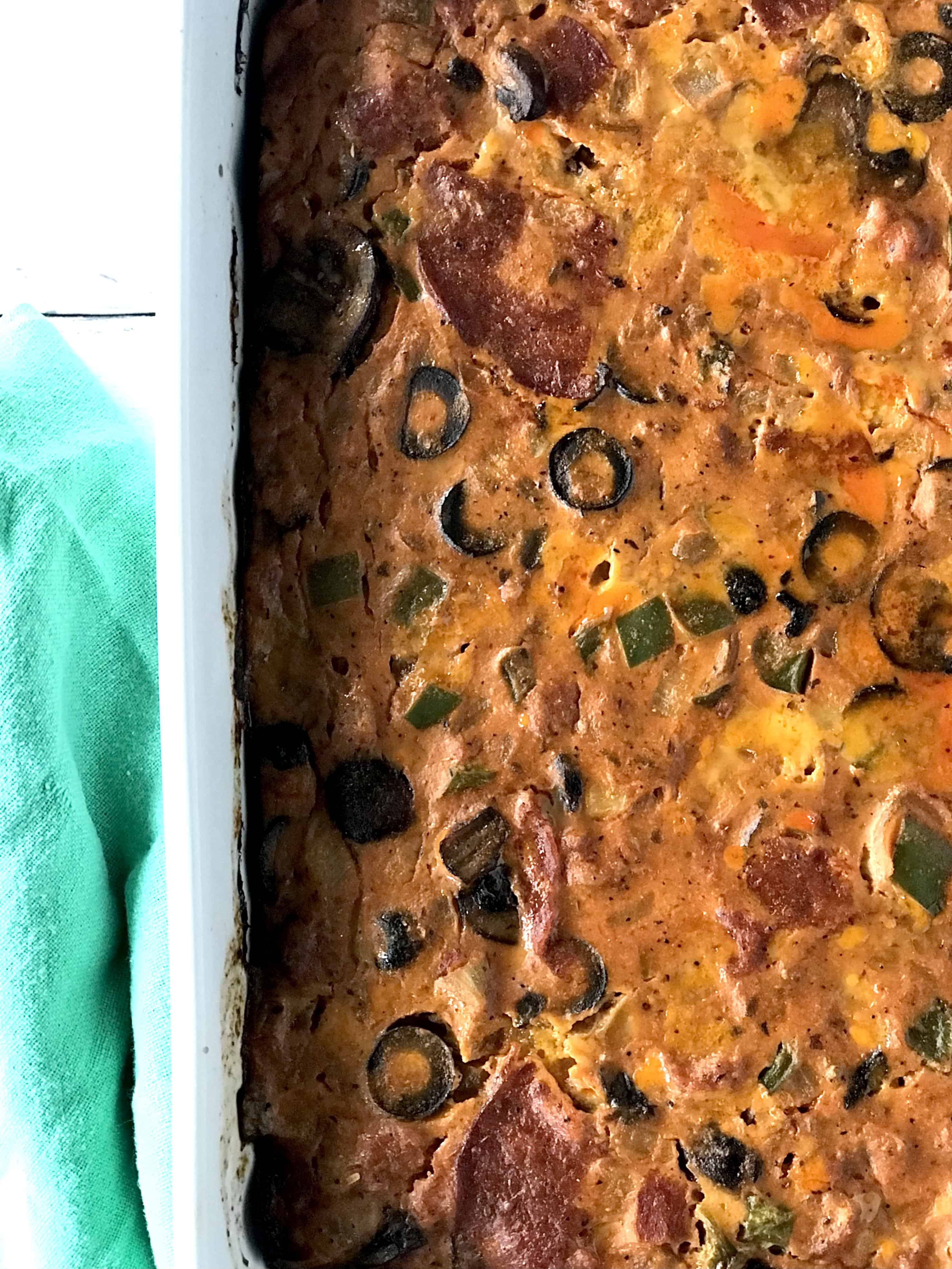 a grain-free casserole with olives and pepperoni in a white baking dish next to a green towel