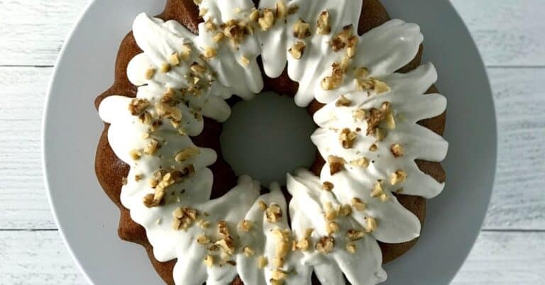 an almond flour cake in a ring shape with a white icing and topped with nuts, sitting on a white cake stand