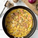 a healthy frittata with smoked salmon, capers and red onion, topped with everything bagel spice mix, sitting in a black skillet on a white wooden table