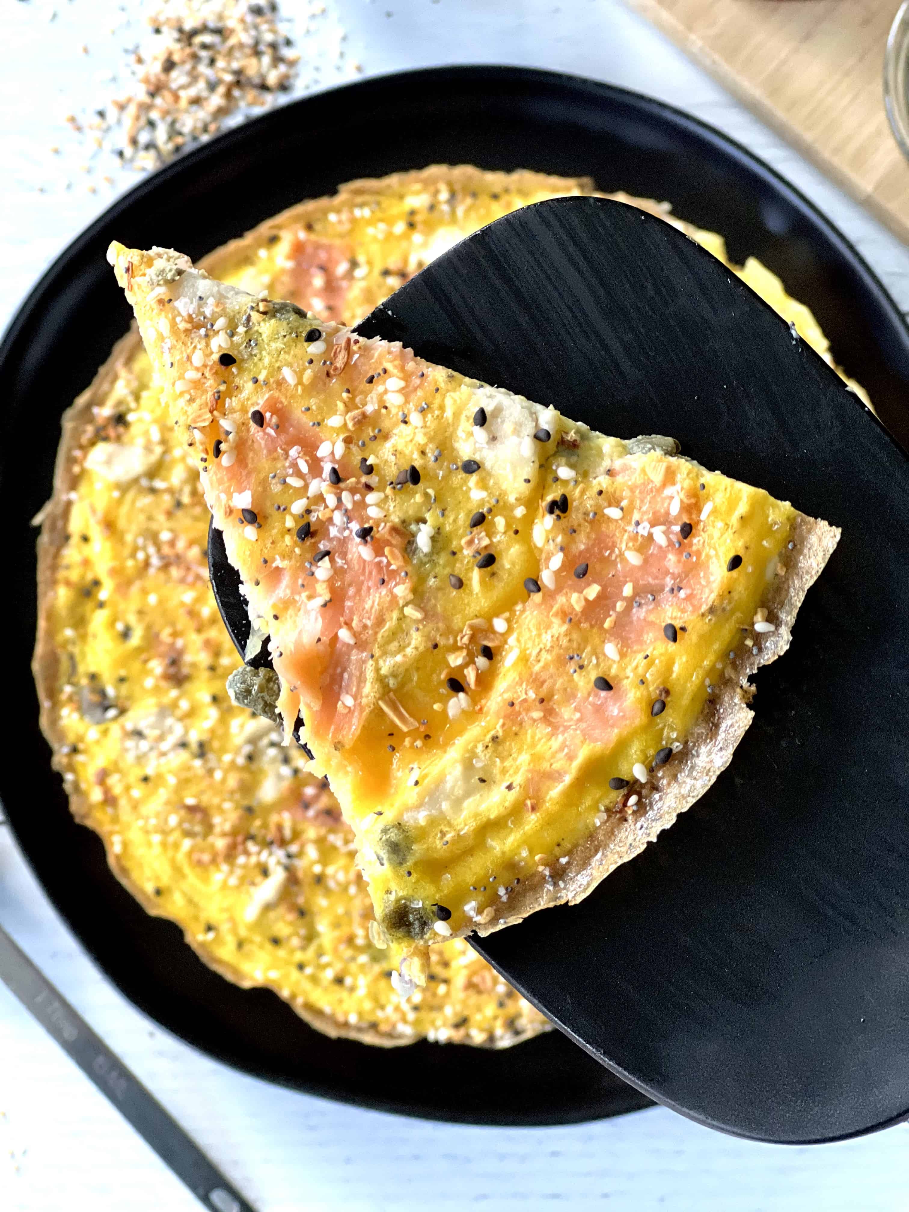 a slice of a cream cheese frittata with smoked salmon on a black spatula, above the whole frittata