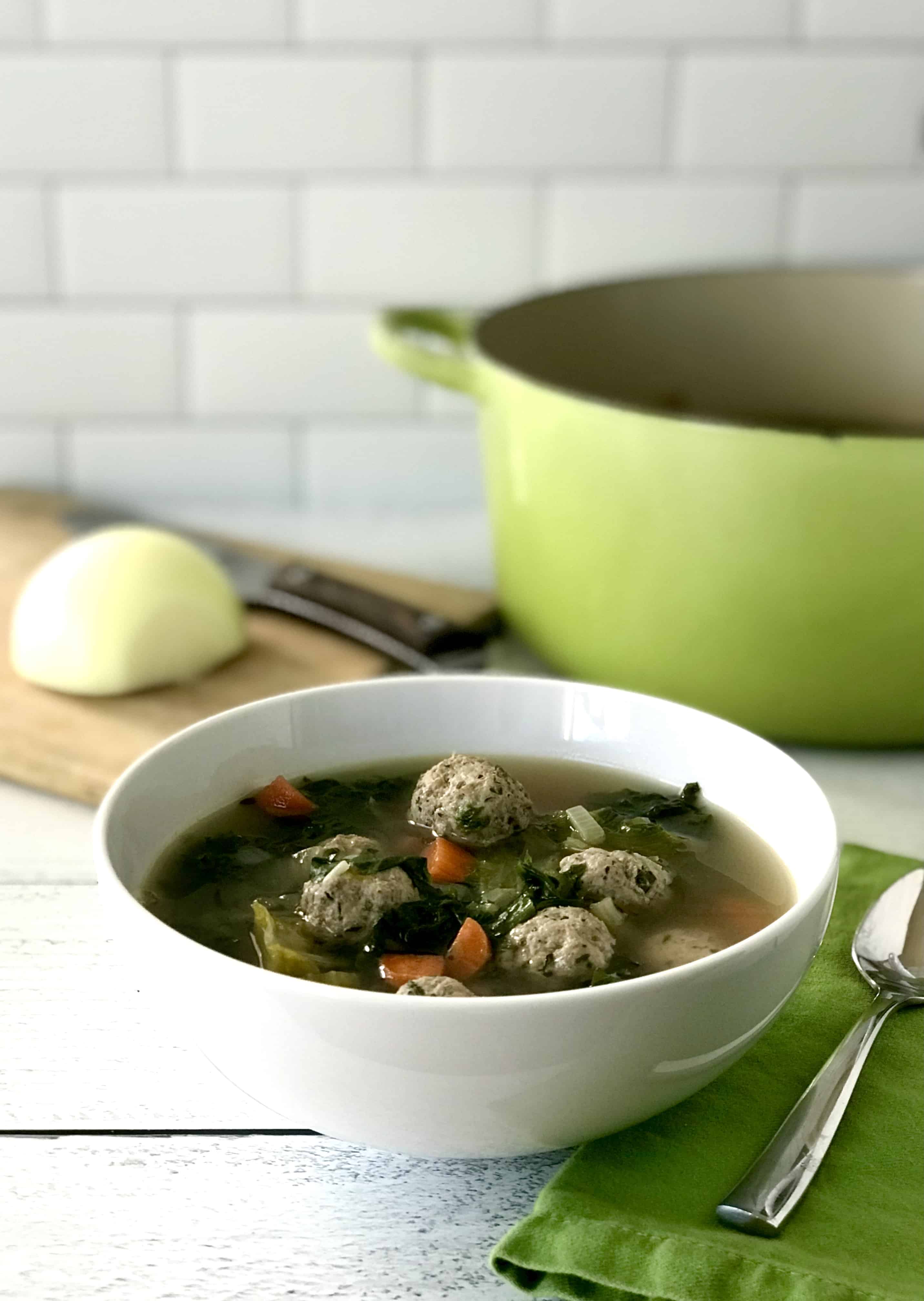 a gluten-free meatball vegetable soup in a white bowl next to a green napkin with a spoon on it