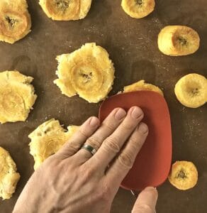 a hand pressing down a spatula turning plantains into tostones
