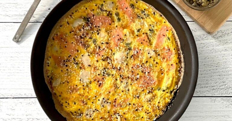 a smoked salmon frittata topped with everything bagel spice mix, in a skillet on a white wooden table