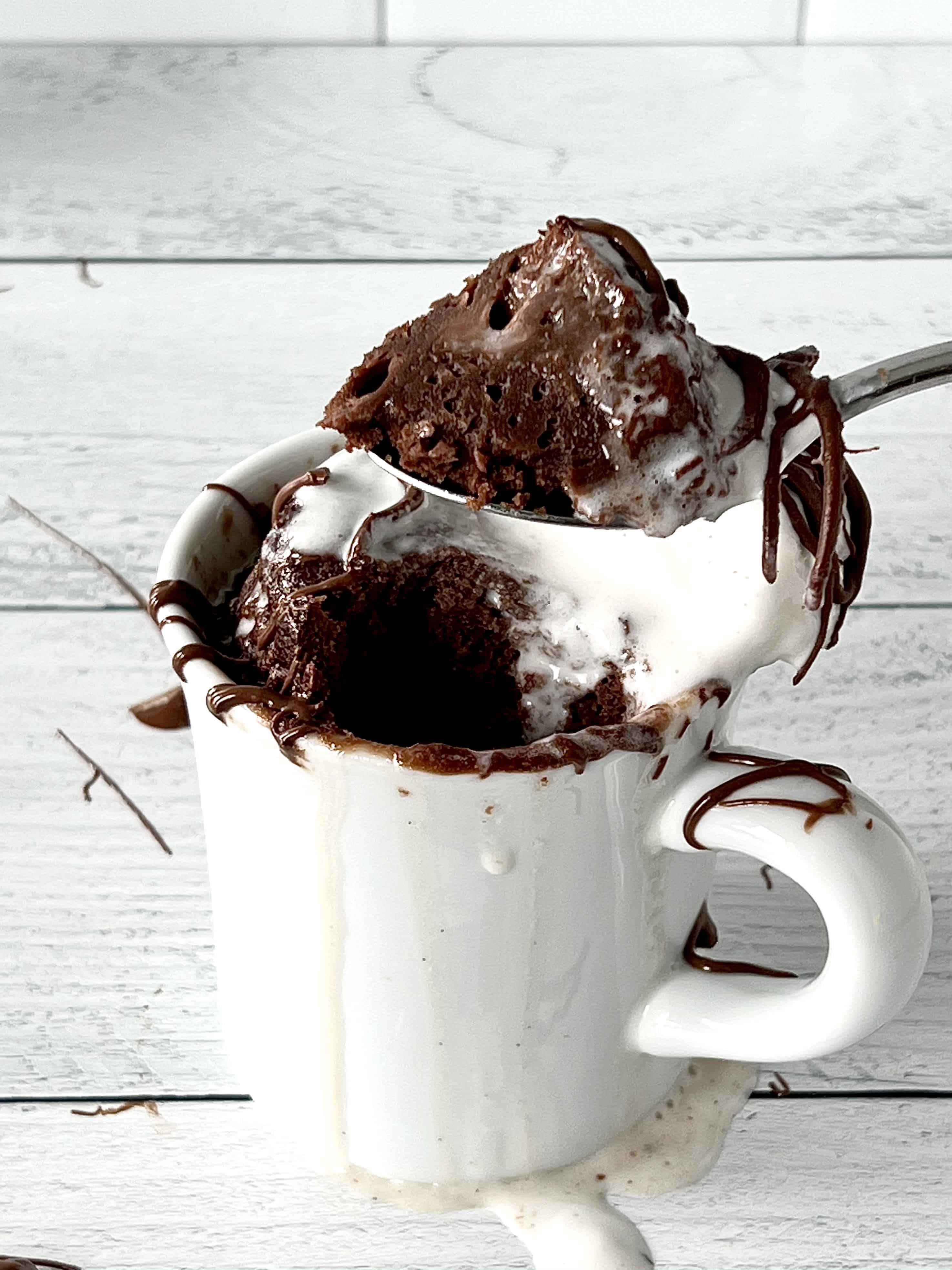 a spoon lifting up a scoop of chocolate hazelnut mug cake over a white mug filled with more of the cake plus vanilla ice cream and drizzles of chocolate hazelnut sauce