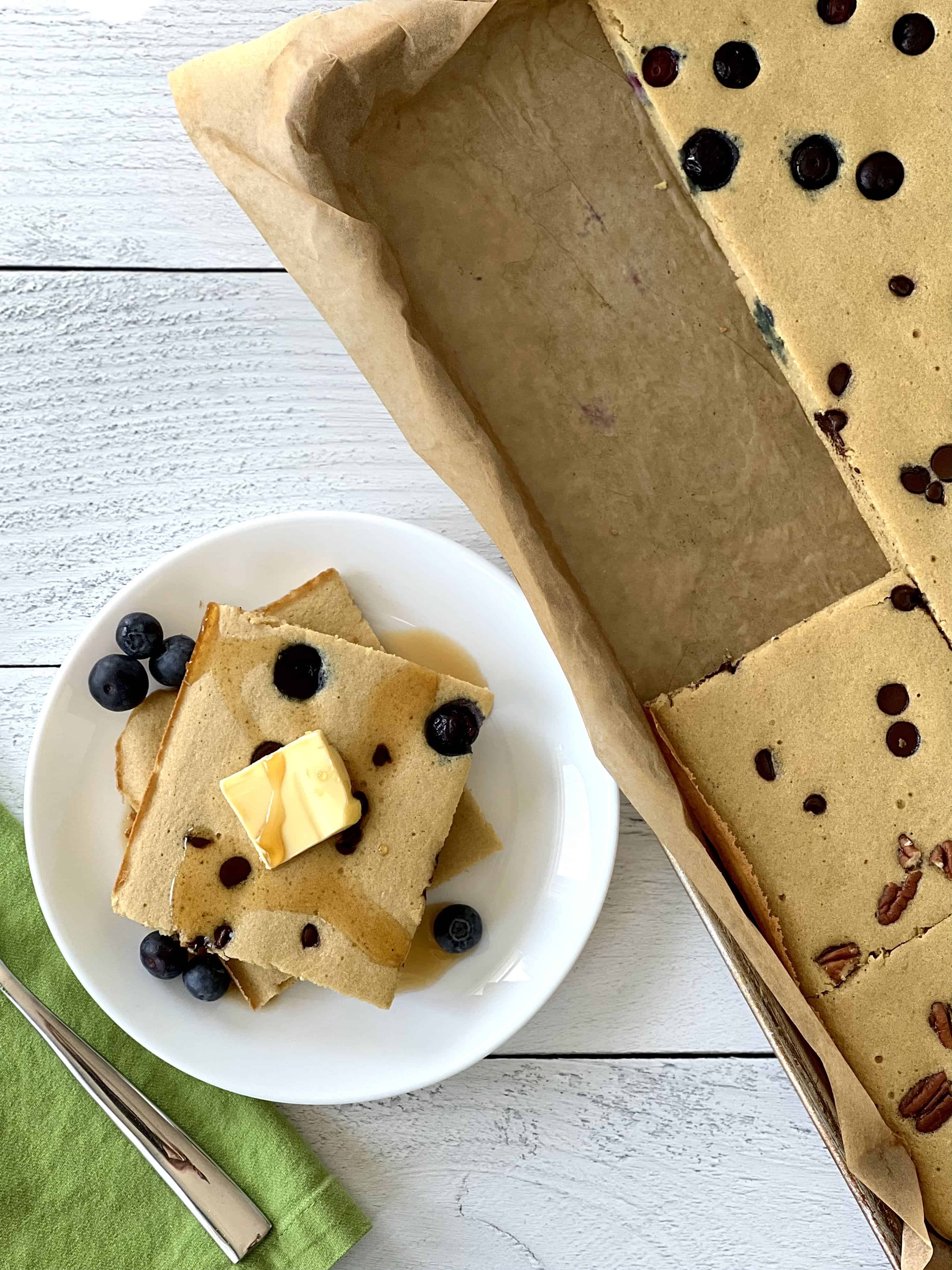 oven baked pancakes in a parchment-lined baking sheet topped with blueberries, chocolate chips and pecans, plus 2 square slices on a white plate topped with butter and maple syrup