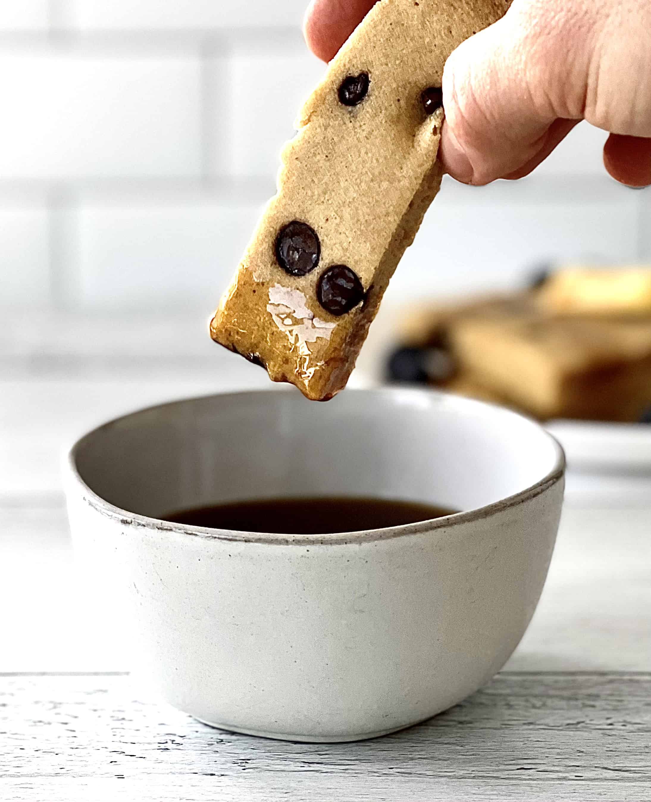 a long rectangular pancake studded with chocolate chips is dipped into a bowl of maple syrup