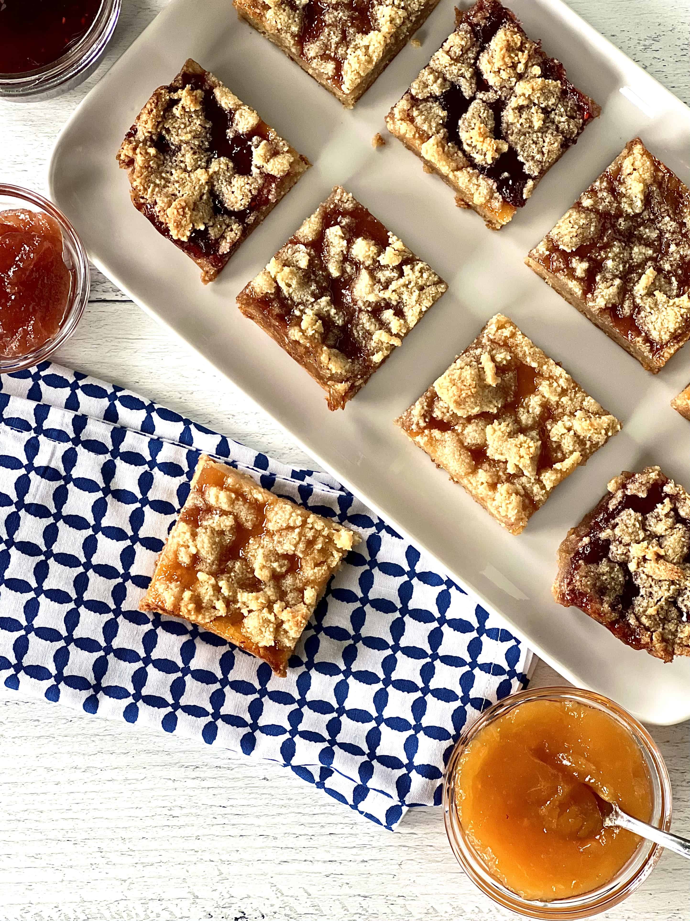healthy crumble bars arranged on a white rectangular platter and one bar on a blue and white patterned napkin next to bowls of jam