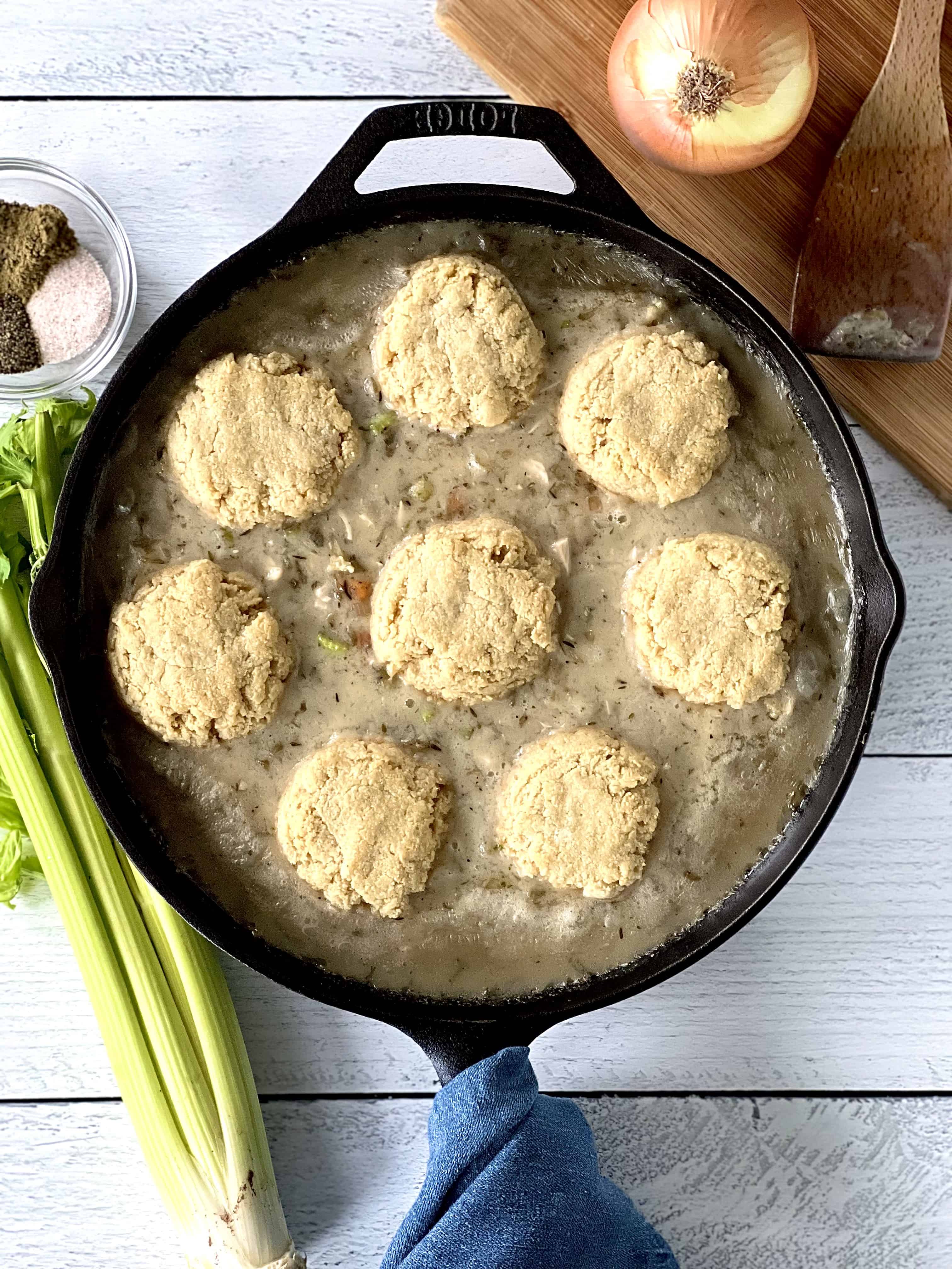 gluten-free biscuits on top of a chicken pot pie filling in a cast iron skillet on a white wooden table next to celery, a bowl of spices and a wooden cutting board with an onion and wooden spoon