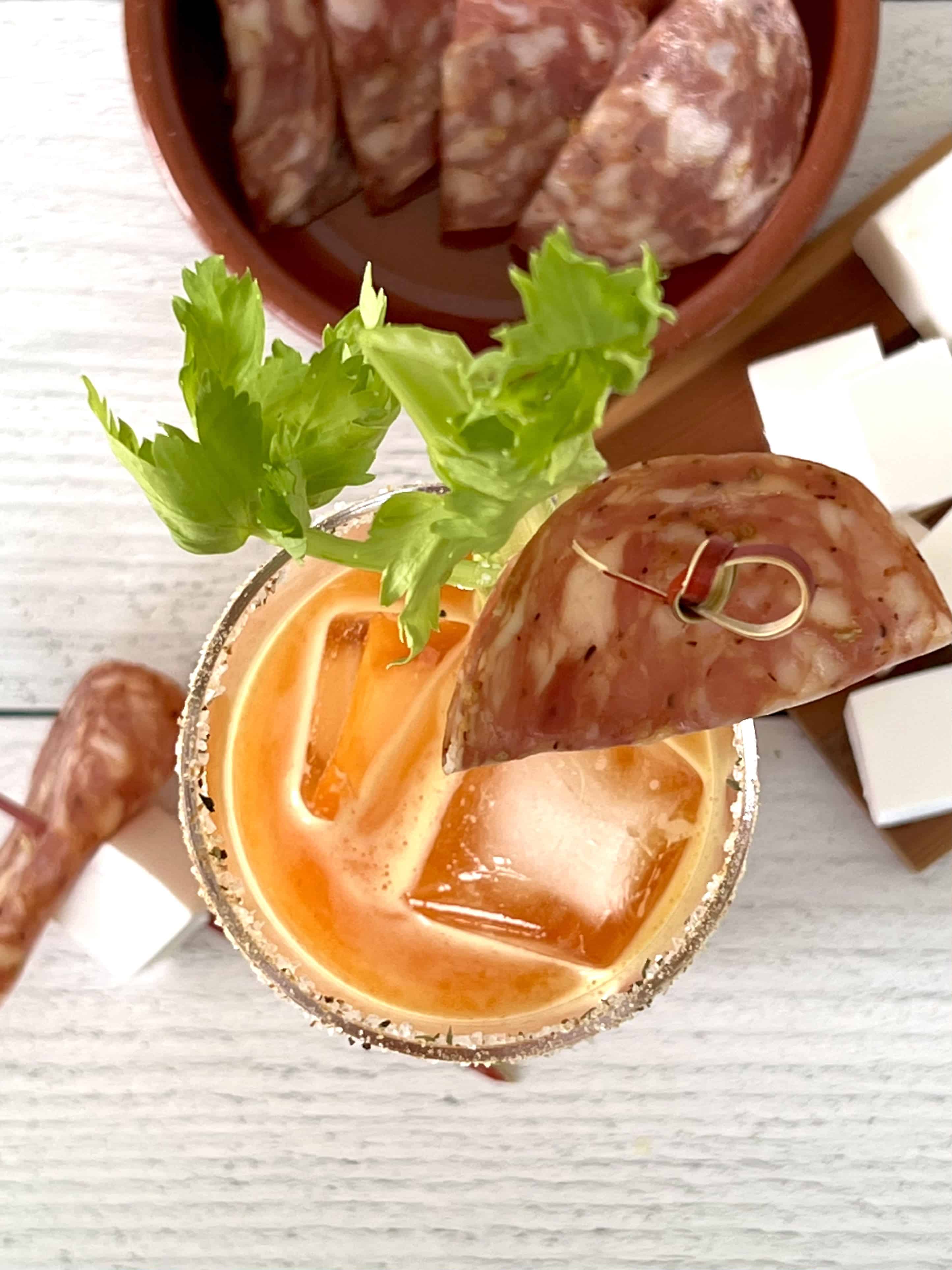 an overhead shot of a carrot vodka cocktail with a salted rim, a celery stick garnish and a toothpick with salami on it