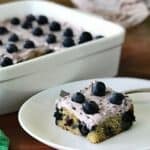 healthy blueberry cake in a white baking dish and a square slice of the cake on a white plate with a fork
