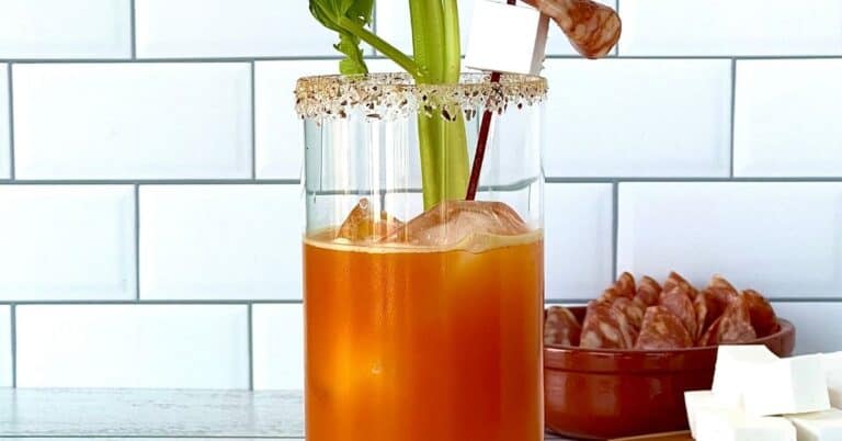 a healthy bloody mary made with carrot juice in a highball glass with a salted rim and a celery stick and salami/feta cube garnish