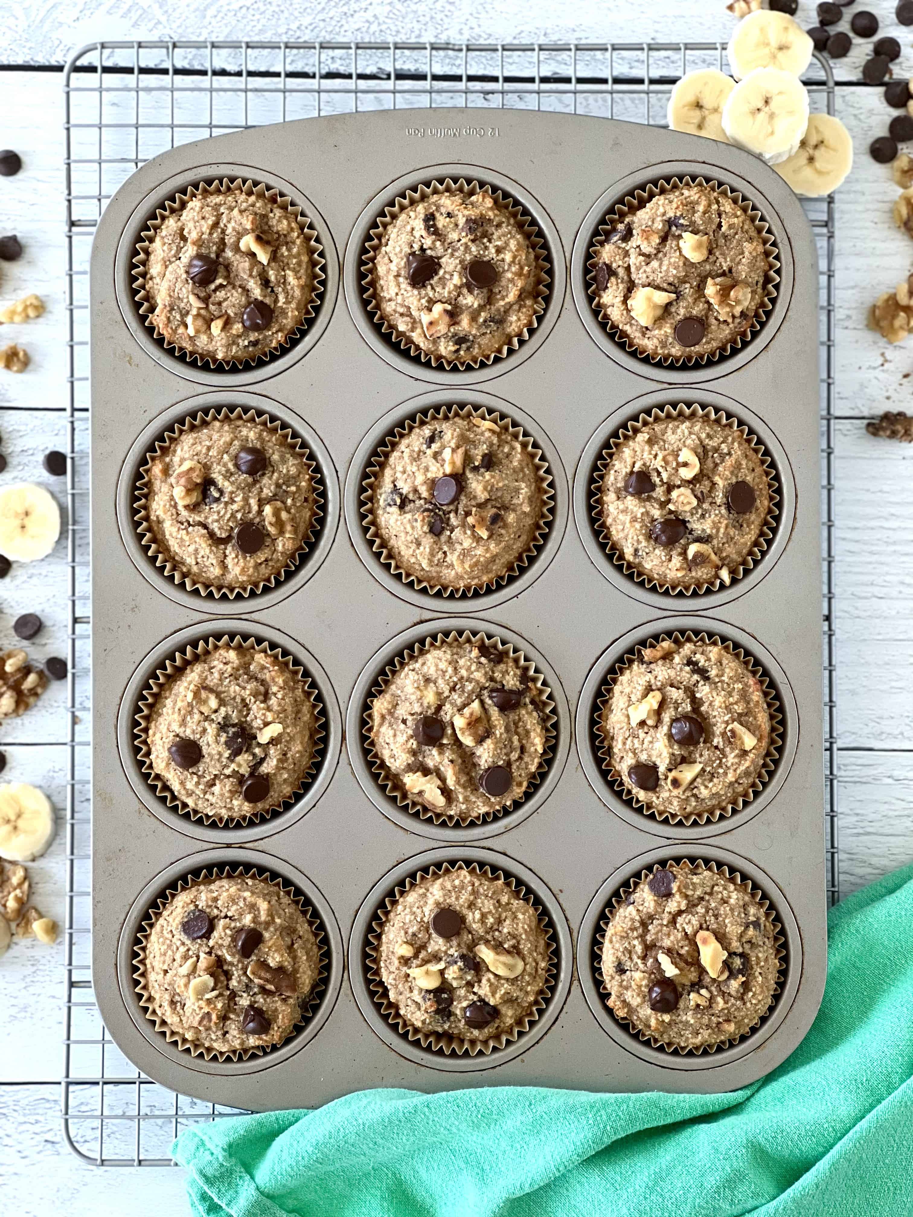 grain-free banana muffins with chocolate chips and walnuts in muffin cups in a muffin pan on a cooling rack on a white wooden table