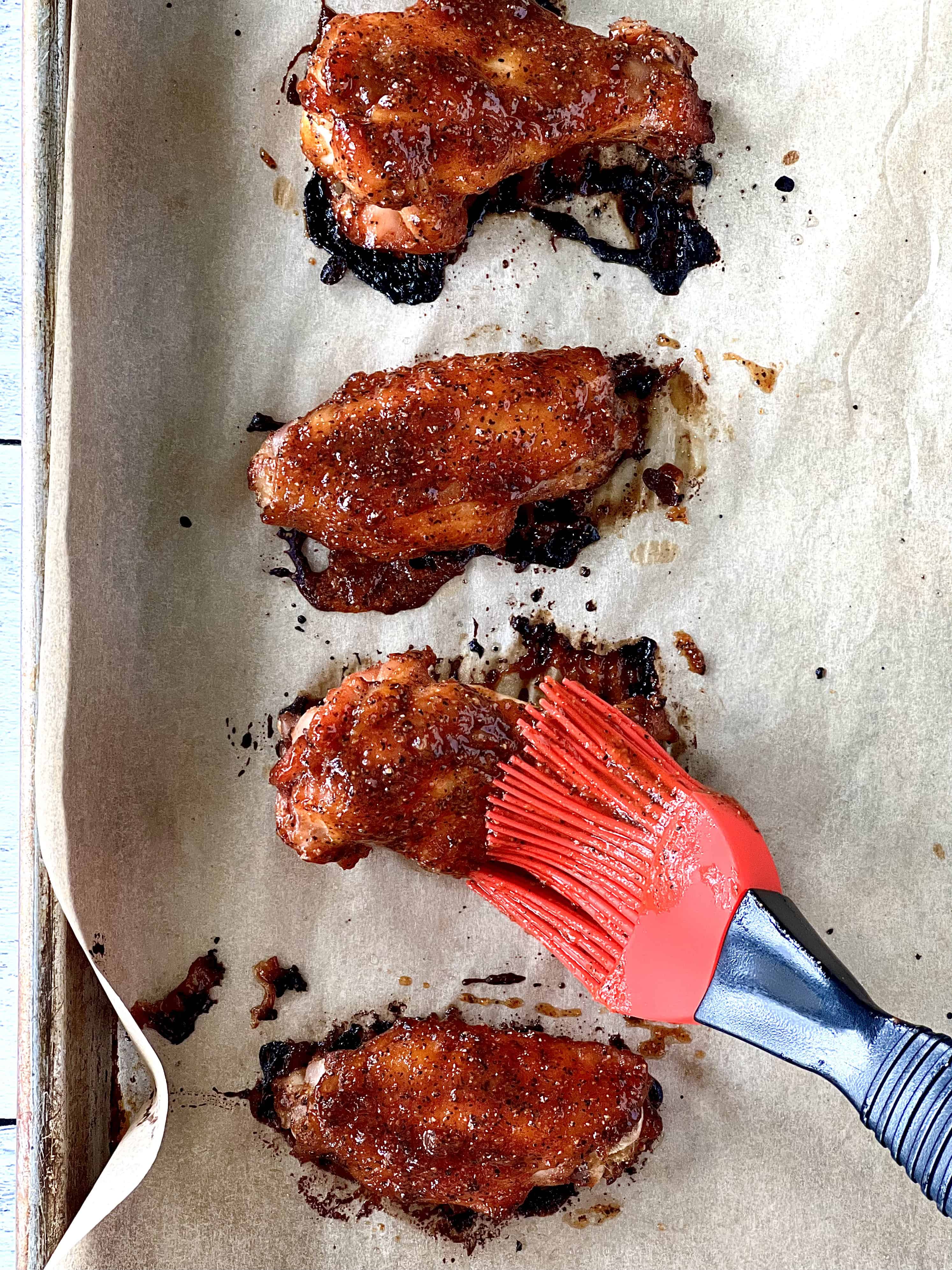 a silicone pastry brush brushing an apricot jam and coconut aminos glaze over chicken wings on a parchment paper-lined baking sheet
