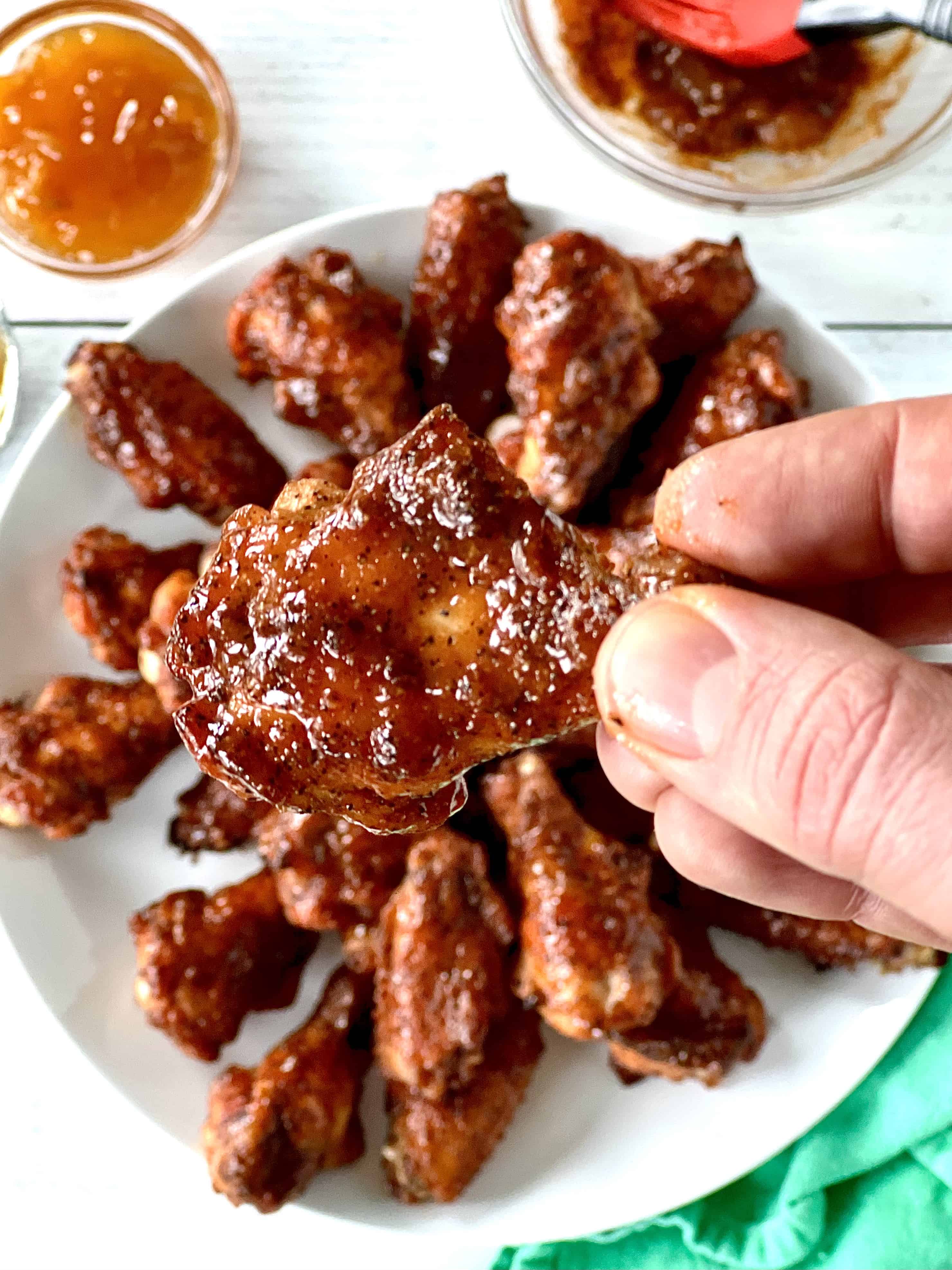 a hand holding up an apricot glazed chicken wing over a white plate of more wings