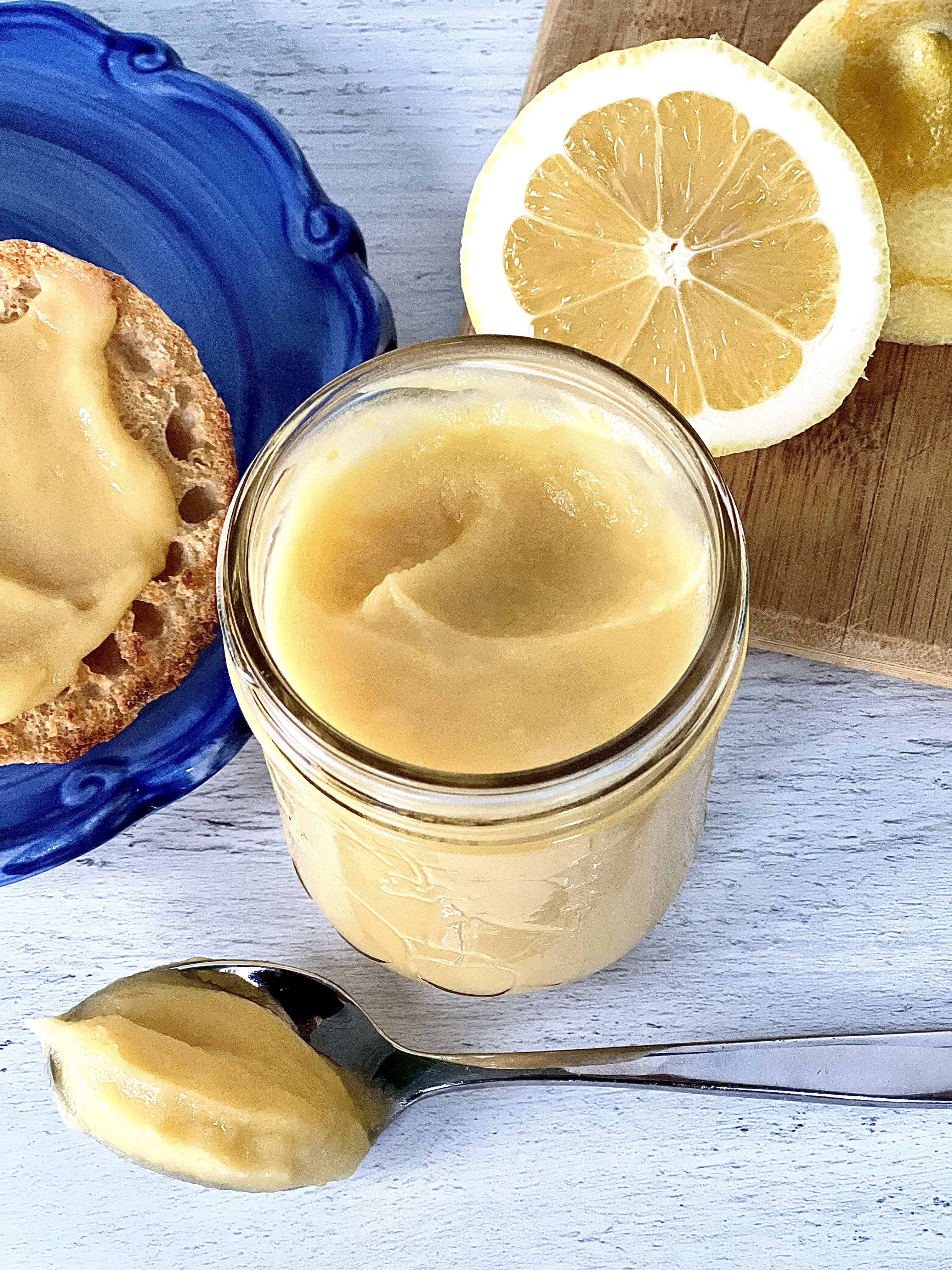 smooth, creamy lemon jam in a mason jar, on a spoon and and two English muffin halves on a blue swirl plate, plus two lemon halves are on a wooden cutting board